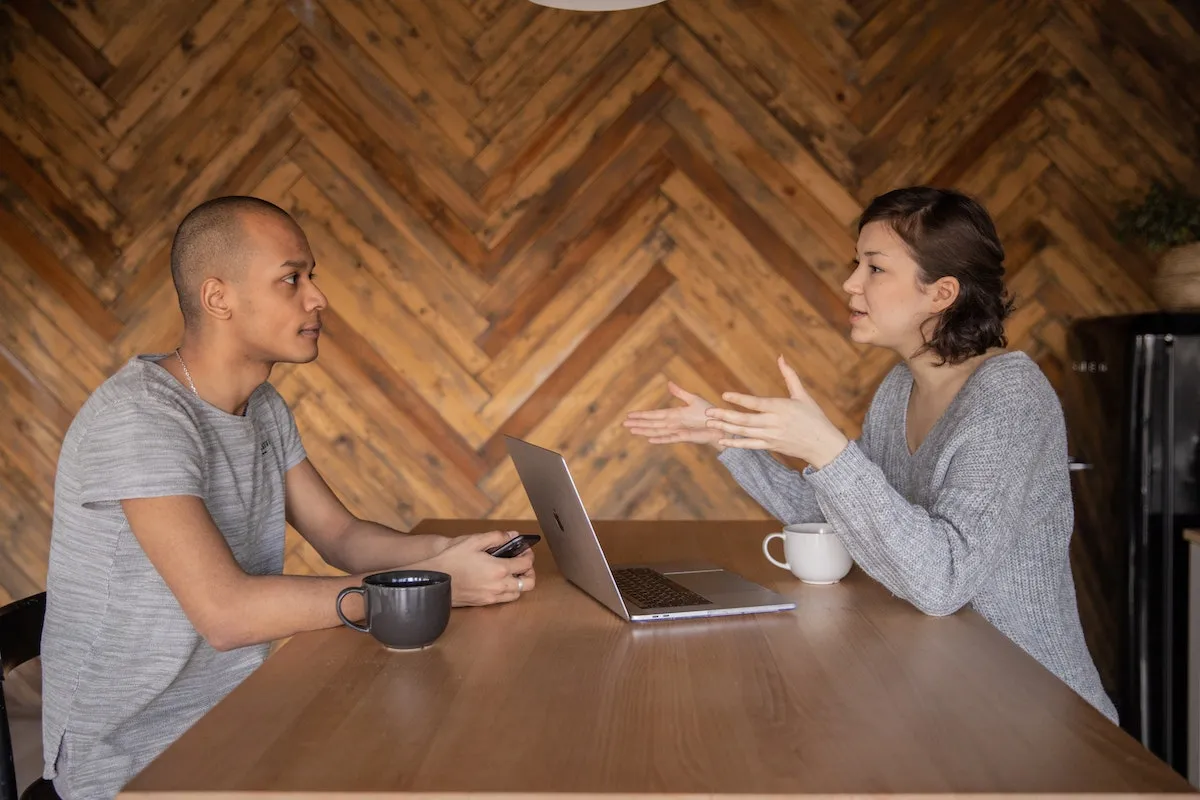 Two colleagues talking across a wooden table with a laptop and coffee mugs